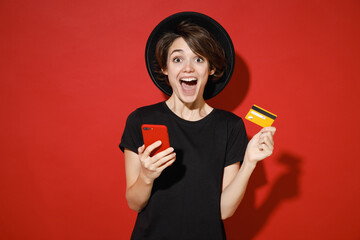 Wall Mural - Surprised young brunette woman 20s years old wearing casual basic black t-shirt hat standing using mobile cell phone hold credit bank card isolated on bright red color background studio portrait.