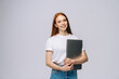 © dikushin - Cheerful young woman student holding laptop computer and looking at camera on isolated gray background. Pretty lady model with red hair emotionally showing facial expressions in studio, copy space.