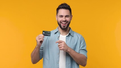 Wall Mural - Cheerful funny bearded young man 20s in casual blue shirt isolated on yellow color background studio. People lifestyle concept. Pointing index finger on credit bank card showing thumb up like gesture