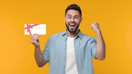 Poster - Happy joyful bearded young man 20s in casual blue shirt isolated on yellow color background studio. People lifestyle concept. Hold gift certificate doing winner gesture celebrating clenching fist