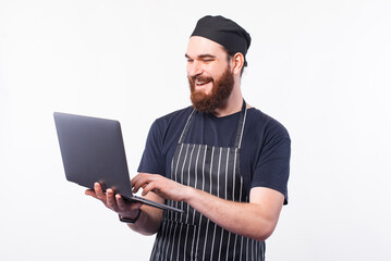 Cheerful chef man in uniform using laptop over white background, looking for recipe.