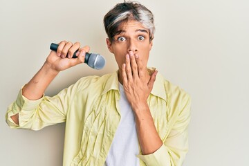 Wall Mural - Young hispanic man singing song using microphone covering mouth with hand, shocked and afraid for mistake. surprised expression