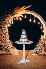 Wall Mural - a white cake on a stand against the backdrop of a wedding acre and Edison lamps on, an evening wedding ceremony at the arch, a cake for guests at a wedding, a birthday cake