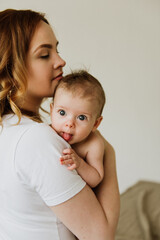 Beautiful mom hugs and kisses the newborn in the studio on a white background. A young mother in a white bodysuit hugs and kisses her newborn daughter in the studio.