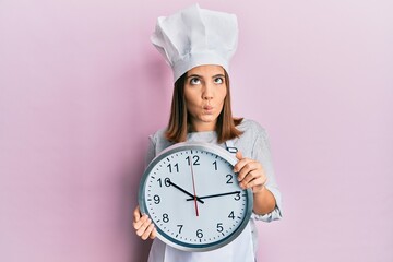 Sticker - Young beautiful woman wearing professional cook uniform and hat holding clock making fish face with mouth and squinting eyes, crazy and comical.