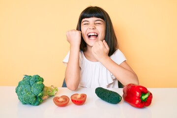 Wall Mural - Young little girl with bang sitting on the table with veggies celebrating surprised and amazed for success with arms raised and eyes closed