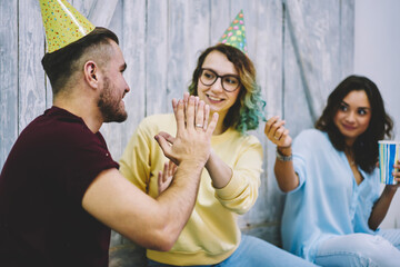 Selective focus on male and female guests of birthday party in cone hats holding hands giving five smiling, angry girl looking jealousy on them feeling envy about boyfriend behaviour on celebration