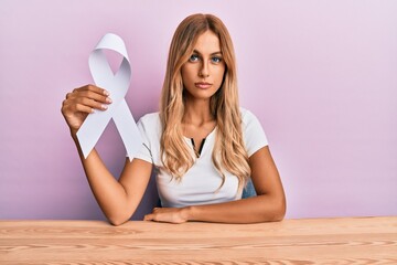 Poster - Beautiful blonde young woman holding white ribbon thinking attitude and sober expression looking self confident