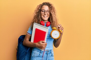 Poster - Beautiful caucasian teenager girl wearing student backpack and holding alarm clock winking looking at the camera with sexy expression, cheerful and happy face.