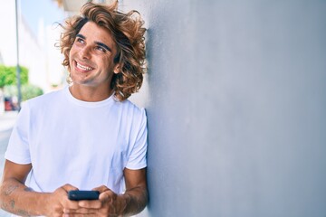 Wall Mural - Young hispanic man smiling happy using smartphone leaning on the wall