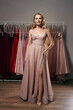 Young beautiful blonde girl wearing a full-length pale pink glitter chiffon draped prom ball gown. Model selecting an outfit for occasion in dress hire service with many options on background.