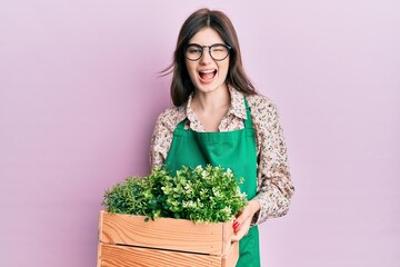 Sticker - Young beautiful caucasian girl wearing gardener apron holding wooden plant pot winking looking at the camera with sexy expression, cheerful and happy face.