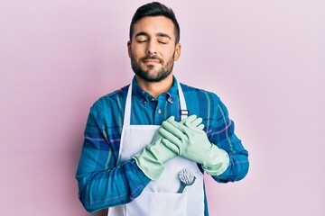 Sticker - Young hispanic man wearing apron smiling with hands on chest with closed eyes and grateful gesture on face. health concept.