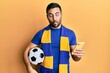 Young hispanic man football hooligan holding soccer ball using smartphone making fish face with mouth and squinting eyes, crazy and comical.