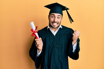Wall Mural - Young hispanic man wearing graduation robe holding diploma screaming proud, celebrating victory and success very excited with raised arm