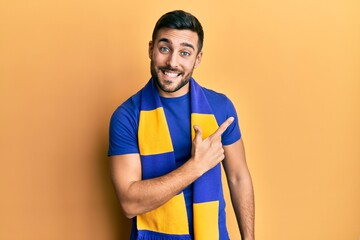 Canvas Print - Young hispanic man football hooligan cheering game smiling cheerful pointing with hand and finger up to the side