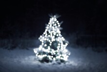 Snow-covered Spruce Tree Illuminated By The Garland Of White Lights Blurred In Bokeh, Close-up. Dark Twilight Evergreen Forest In The Background. Winter Landscape. Christmas Celebration, Decoration