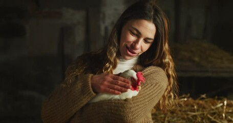 Wall Mural - Cinematic shot of young happy female farmer is caressing with love and care ecologically grown white hen for biological genuine food products industry in hay barn of countryside agricultural farm.