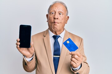 Wall Mural - Senior caucasian man holding smartphone and credit card making fish face with mouth and squinting eyes, crazy and comical.