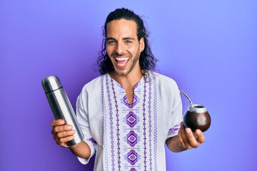 Poster - Young handsome man with long hair drinking mate infusion smiling and laughing hard out loud because funny crazy joke.