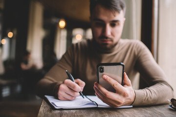 Modern smartphone in hand of concentrated man taking notes in planner