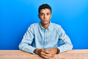 Poster - Young handsome african american man wearing casual clothes sitting on the table relaxed with serious expression on face. simple and natural looking at the camera.