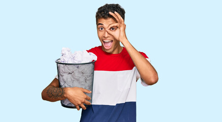 Wall Mural - Young handsome african american man holding paper bin full of crumpled papers smiling happy doing ok sign with hand on eye looking through fingers