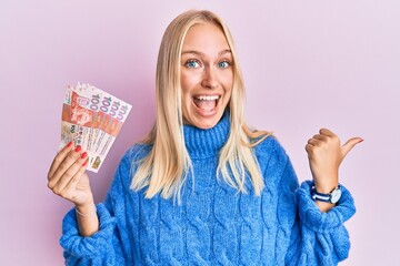 Poster - Young blonde girl holding hong kong 10 dollars banknotes pointing thumb up to the side smiling happy with open mouth