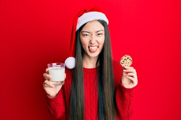 Sticker - Young chinese woman wearing christmas hat holding cookies and milk sticking tongue out happy with funny expression.