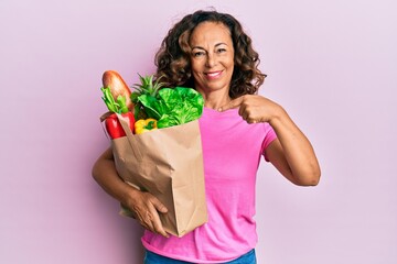 Middle age hispanic woman holding paper bag with bread and groceries smiling happy pointing with hand and finger