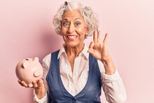 Senior Grey-haired Woman Holding Piggy Bank Doing Ok Sign With Fingers, Smiling Friendly Gesturing Excellent Symbol