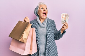 Wall Mural - Senior grey-haired woman holding shopping bags and uk pounds banknotes angry and mad screaming frustrated and furious, shouting with anger. rage and aggressive concept.