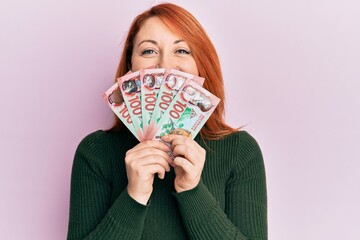 Poster - Beautiful redhead woman holding 100 new zealand dollars banknote smiling with a happy and cool smile on face. showing teeth.