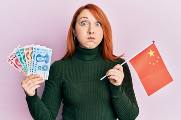 Sticker - Beautiful redhead woman holding yuan chinese banknotes and china flag puffing cheeks with funny face. mouth inflated with air, catching air.