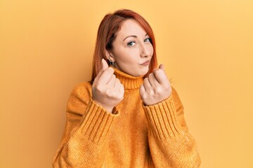 Poster - Beautiful redhead woman wearing casual winter sweater over yellow background doing money gesture with hands, asking for salary payment, millionaire business