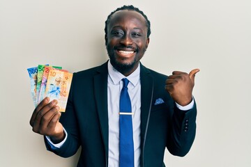 Poster - Handsome young black man wearing business suit holding franc swiss banknotes pointing thumb up to the side smiling happy with open mouth