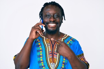 Sticker - Young african american man with braids wearing ethnic tshirt having conversation talking on the smartphone smiling happy pointing with hand and finger