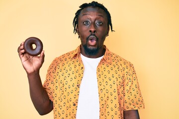 Sticker - Young african american man with braids holding donut scared and amazed with open mouth for surprise, disbelief face