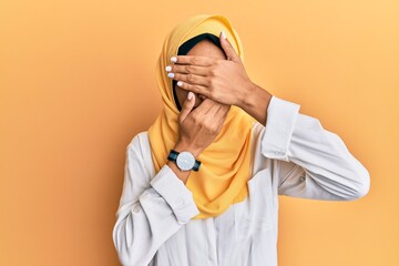 Wall Mural - Young brunette arab woman wearing traditional islamic hijab scarf covering eyes and mouth with hands, surprised and shocked. hiding emotion