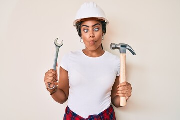 Poster - Young african american woman with braids wearing hardhat holding hammer and wrench making fish face with mouth and squinting eyes, crazy and comical.