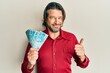 Middle age handsome man holding 100 brazilian real banknotes smiling happy and positive, thumb up doing excellent and approval sign