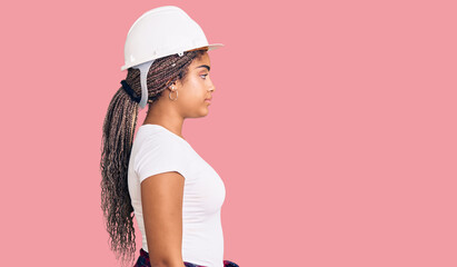 Canvas Print - Young african american woman with braids wearing hardhat and builder clothes looking to side, relax profile pose with natural face with confident smile.