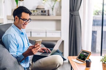 Wall Mural - Young smiling asian man relaxing using laptop computer working and video conference meeting at home.Young creative man looking at screen typing message with smartphone.work from home concept