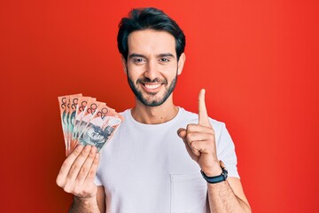 Sticker - Young hispanic man holding australian dollars smiling with an idea or question pointing finger with happy face, number one