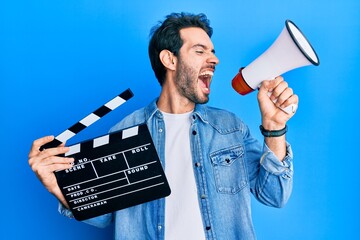 Sticker - Young hispanic man holding video film clapboard and megaphone angry and mad screaming frustrated and furious, shouting with anger. rage and aggressive concept.