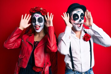 Wall Mural - Couple wearing day of the dead costume over red smiling cheerful playing peek a boo with hands showing face. surprised and exited