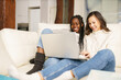 © javiindy - Two female student friends sitting on the couch at home using a laptop.