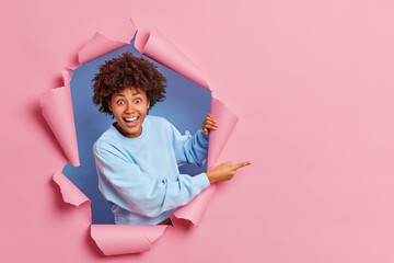 Wall Mural - Cheerful amazed dark skinned Afro American woman indicates at blank space demonstrates advertisement wears blue jumper has happy expression breaks through paper background. Promotion concept