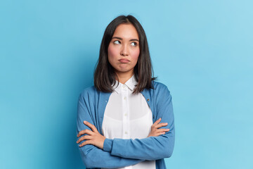 Wall Mural - Portrait of displeased offended young Asian woman with dark hair keeps arms folded looks angrily aside doesnt agree with somebodys opinion wears neat clothes isolated over blue studio background