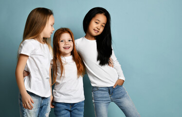 Mixed race international happy girls friends in jeans and white t-shirts standing and hugging together smiling laughing over light blue background, copy space. Happy childhood, stylish look